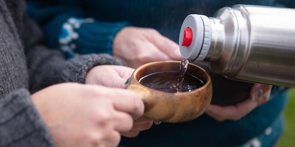 Lähikuvassa henkilö kaataa toisen henkilön puiseen kuksaan vettä termospullosta.